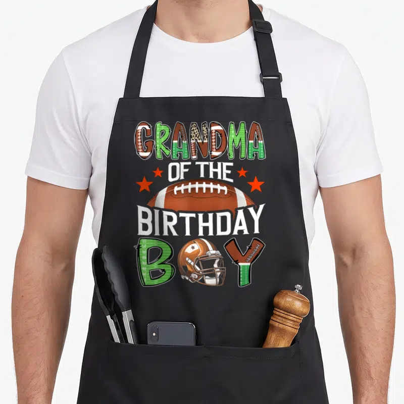 Game grandma aprons - Grandma of the Birthday Boy text with football and helmet graphic in brown and green — shown on a black cotton canvas apron against a white background