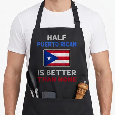 Puerto half birthday apron - Centered Puerto Rican flag with text HALF PUERTO RICAN IS BETTER THAN NONE in red, white — shown on a black cotton canvas apron against a white background