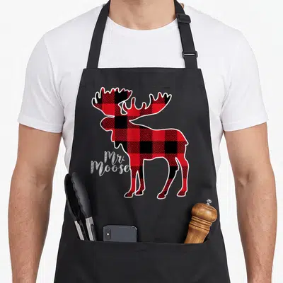 Family red plaid apron - Cartoon moose filled with red black plaid pattern and white Mr. Moose script text — shown on a black cotton canvas apron against a white background