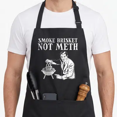 Brisket men's barbecue apron - White text SMOKE BRISKET NOT METH above a grey suited man grilling meat on a kettle grill — shown on a black cotton canvas apron against a white background