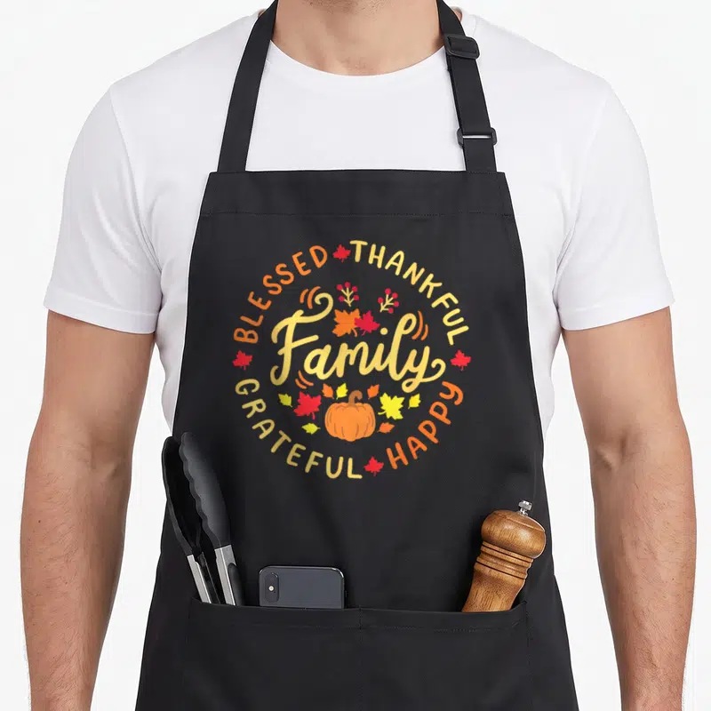 Blessed family thankful apron - Cartoon pumpkin and circular text BLESSED THANKFUL Family GRATEFUL HAPPY in autumn — shown on a black cotton canvas apron against a white background