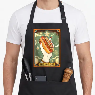 Bun hot dog apron - Retro tarot card style hand holding hot dog with mustard and THE HOT DOG text — shown on a black cotton canvas apron against a white background