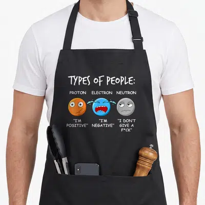 Physicist teacher apron - Orange proton, blue electron, grey neutron cartoon with physics pun quotes — shown on a black cotton canvas apron against a white background
