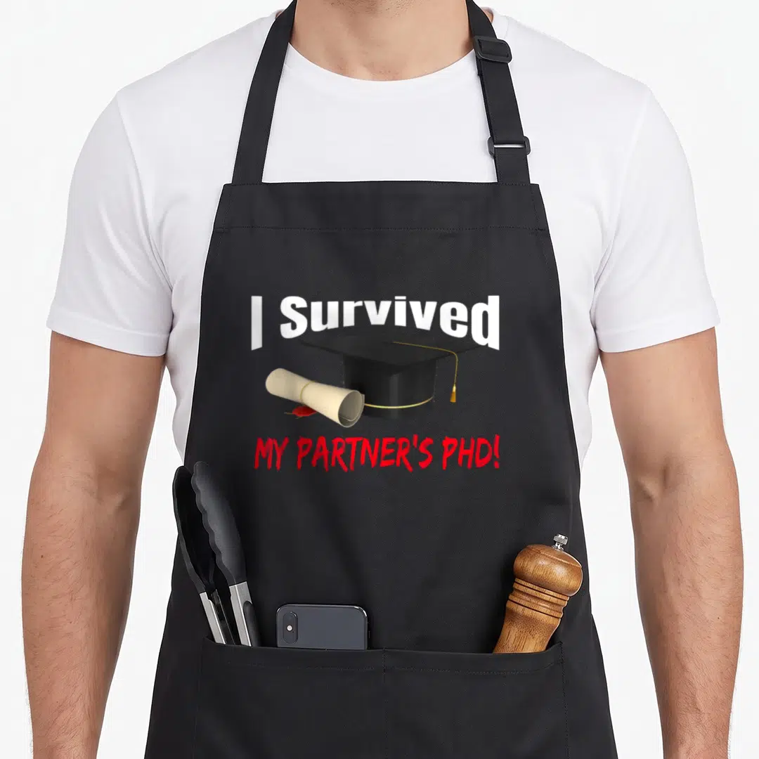 Survived funny graduation apron - I Survived My Partner's PhD text with black cap and cream diploma illustration — shown on a black cotton canvas apron against a white background