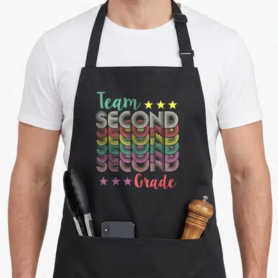 Teacher school 2nd apron - Retro rainbow text stacking the word SECOND seven times between Team and Grade with yellow — shown on a black cotton canvas apron against a white background