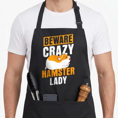 Funny beware crazy hamster lady apron - Cartoon hamster in orange and white with stacked text BEWARE CRAZY HAMSTER LADY — shown on a black cotton canvas apron against a white background