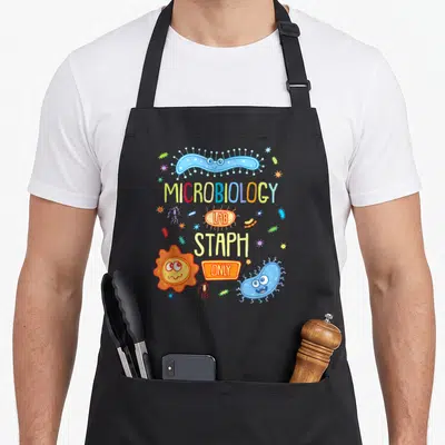 Petri microbiologist apron - Cartoon blue and orange bacteria surrounding MICROBIOLOGY LAB STAPH ONLY text — shown on a black cotton canvas apron against a white background
