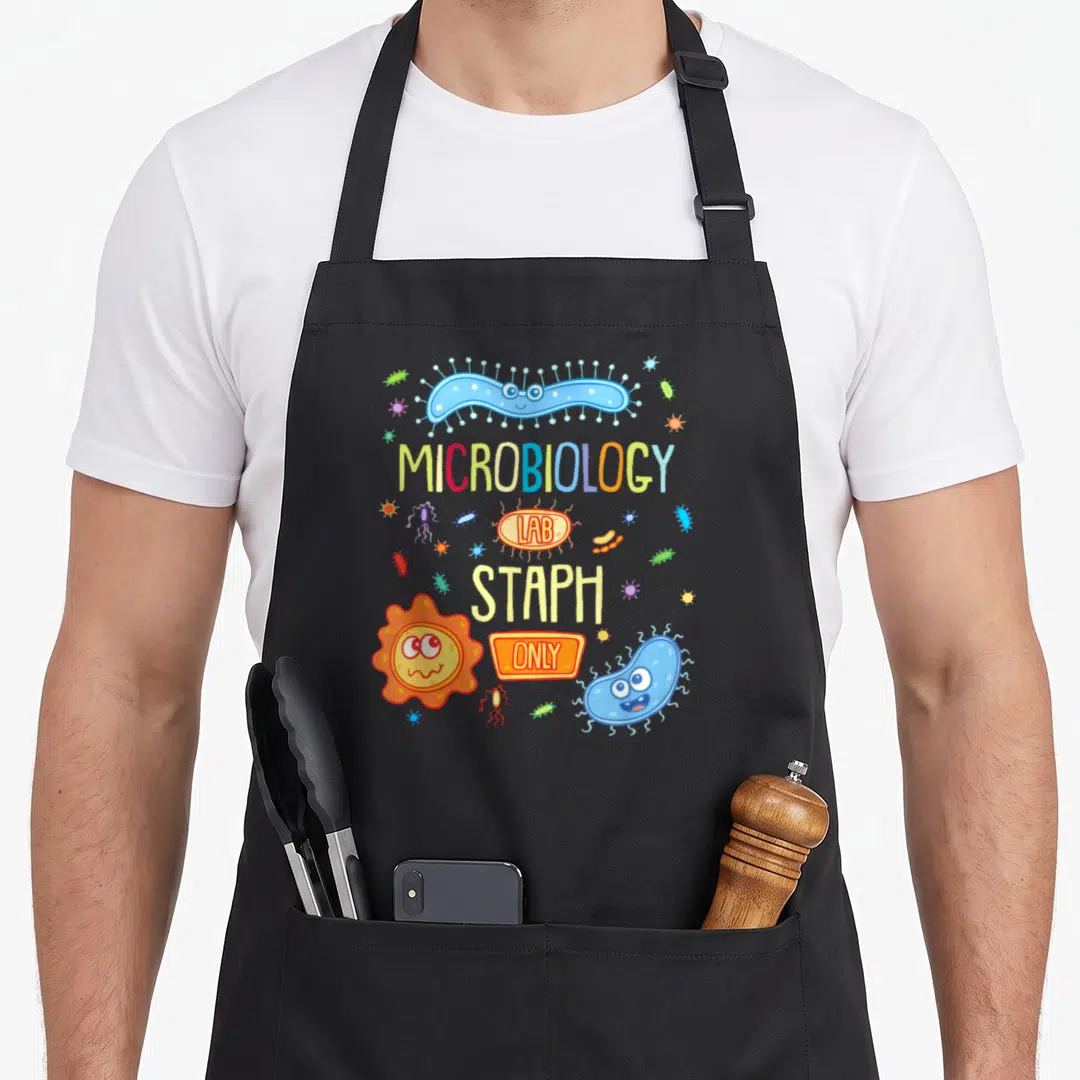 Petri microbiologist apron - Cartoon blue and orange bacteria surrounding MICROBIOLOGY LAB STAPH ONLY text — shown on a black cotton canvas apron against a white background