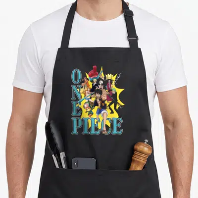 Burst collage anime character apron - One Piece text and Monkey D Luffy group art on yellow starburst — shown on a black cotton canvas apron against a white background