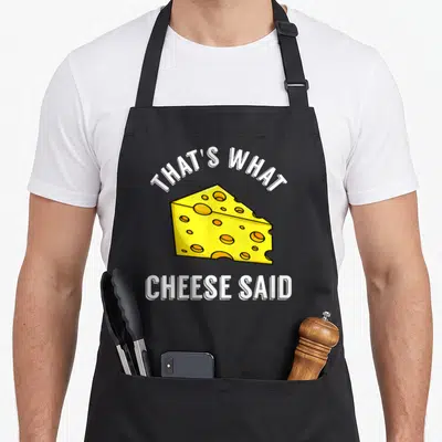 Cheese cheesy aprons - Yellow cartoon cheese wedge with white text That's What Cheese Said — shown on a black cotton canvas apron against a white background