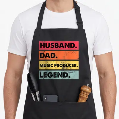 Husband making aprons - Stacked red orange yellow blue bars with black text Husband Dad Music Producer Legend distressed — shown on a black cotton canvas apron against a white background
