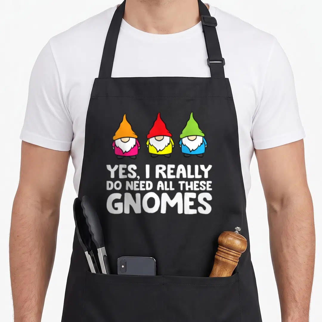 Gnome do i need an apron - Three cartoon gnomes with orange red and green hats standing over white stacked text — shown on a black cotton canvas apron against a white background
