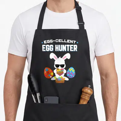 Cellent easter egg apron - Cartoon bunny with sunglasses holding egg under EGG-CELLENT EGG HUNTER text with colored eggs — shown on a black cotton canvas apron against a white background