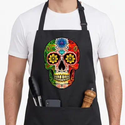 Muerto sugar skull apron - Cartoon sugar skull with blue flower, diamond forehead, and yellow eye flowers in green — shown on a black cotton canvas apron against a white background