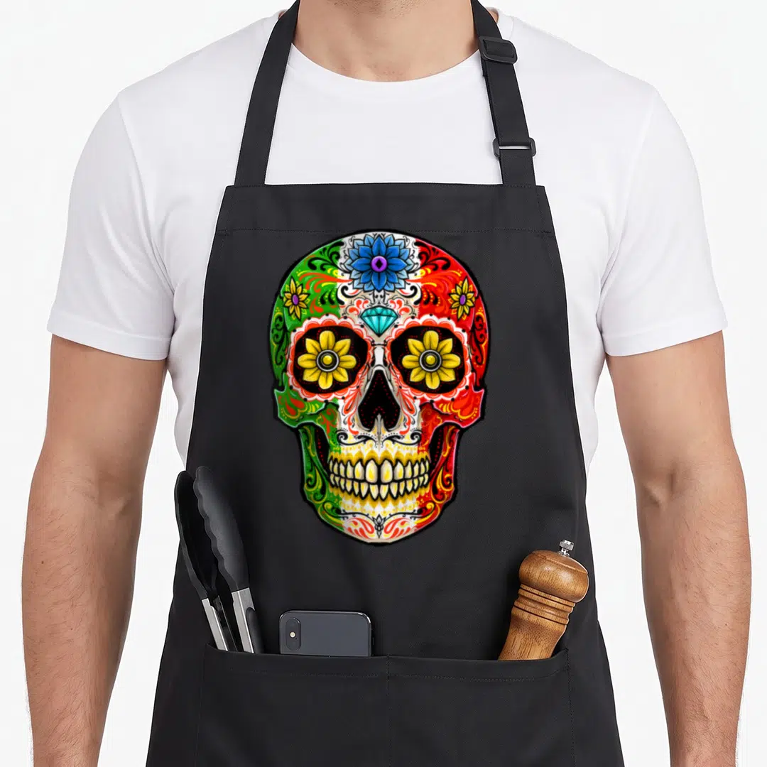 Muerto sugar skull apron - Cartoon sugar skull with blue flower, diamond forehead, and yellow eye flowers in green — shown on a black cotton canvas apron against a white background