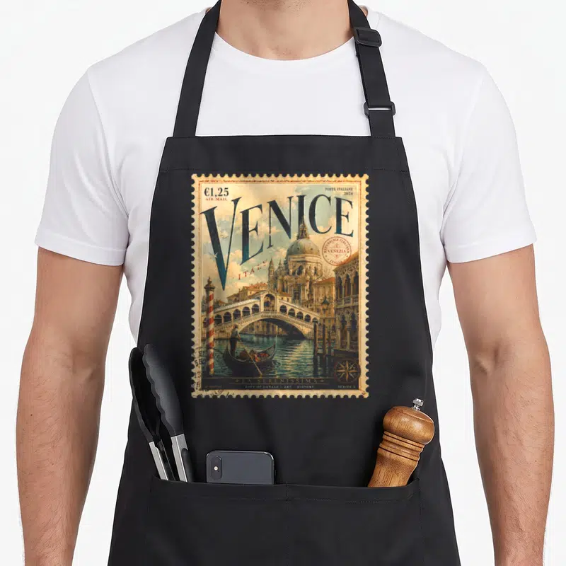 Postage italy apron - Retro Venice stamp design with blue VENICE text, red stamp, and gondola scene on tan background — shown on a black cotton canvas apron against a white background