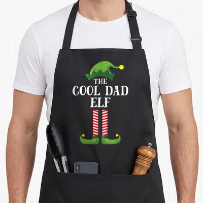 Elf personalized family aprons - Green elf hat and red striped legs framing white text on black apron — shown on a black cotton canvas apron against a white background