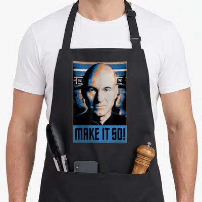 Generation novelty apron - Stylized portrait of a man above blue block reading MAKE IT SO in orange and black — shown on a black cotton canvas apron against a white background