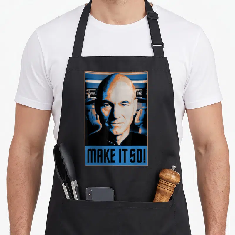 Generation novelty apron - Stylized portrait of a man above blue block reading MAKE IT SO in orange and black — shown on a black cotton canvas apron against a white background