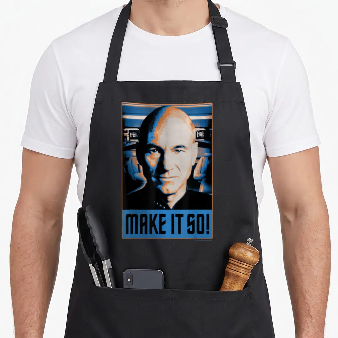 Generation novelty apron - Stylized portrait of a man above blue block reading MAKE IT SO in orange and black — shown on a black cotton canvas apron against a white background