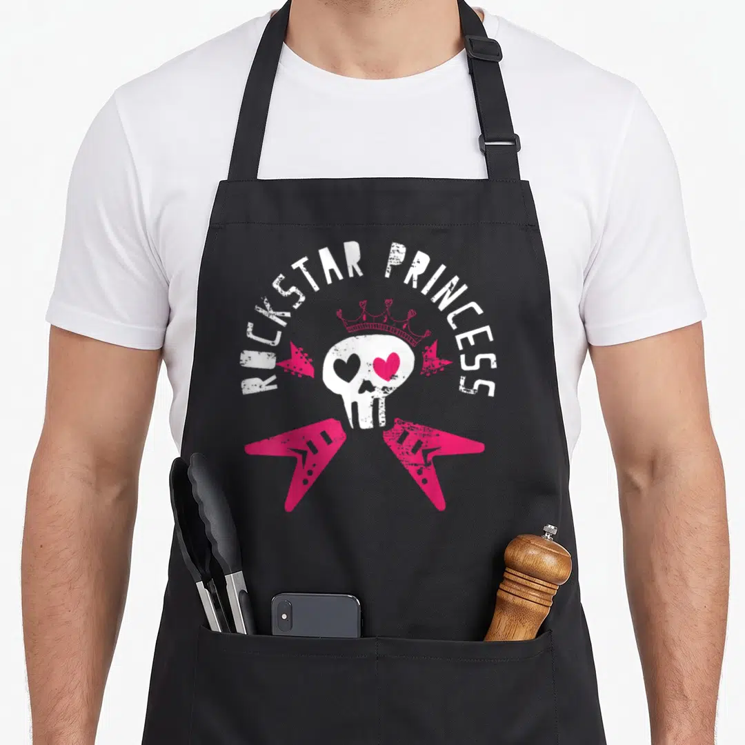 Rockstar princess apron - White skull with pink crown and crossed pink guitars under arched text — shown on a black cotton canvas apron against a white background