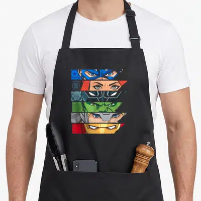 Eye super hero aprons - Six horizontal strips of superhero eyes in blue, red, green, gold, and silver cartoon art — shown on a black cotton canvas apron against a white background