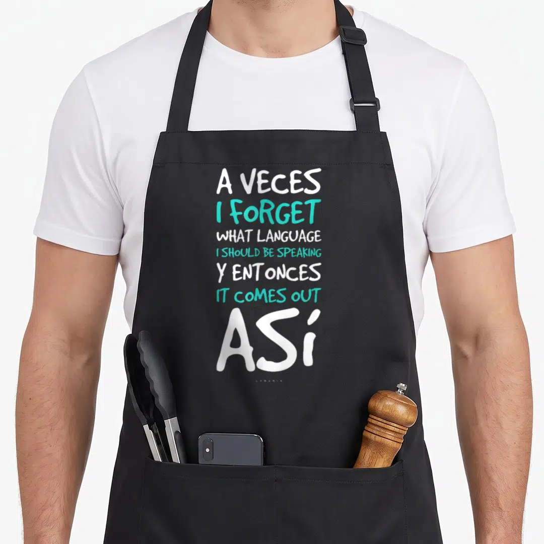 Vece spanish aprons - Stacked white and teal text reading A Veces I Forget What Language Y Entonces It Comes Out Asi — shown on a black cotton canvas apron against a white background