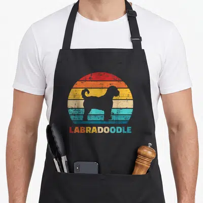 Labradoodle havanese apron - Grey dog silhouette on red orange yellow teal striped sun with labradoodle text — shown on a black cotton canvas apron against a white background