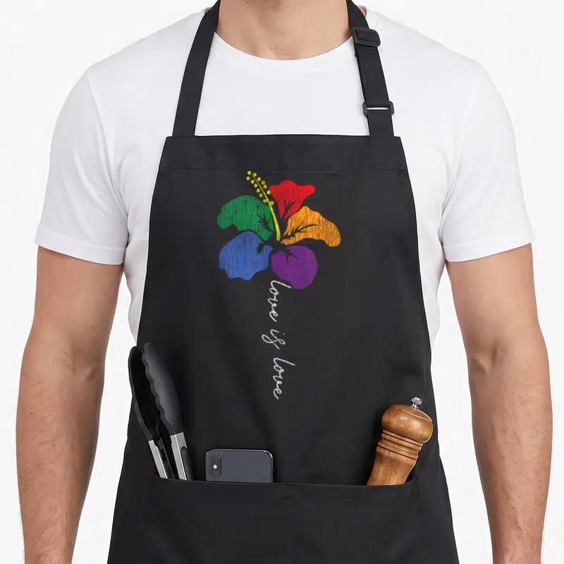 Rainbow white lily apron - Rainbow hibiscus flower with vertical love is love text in watercolor style — shown on a black cotton canvas apron against a white background