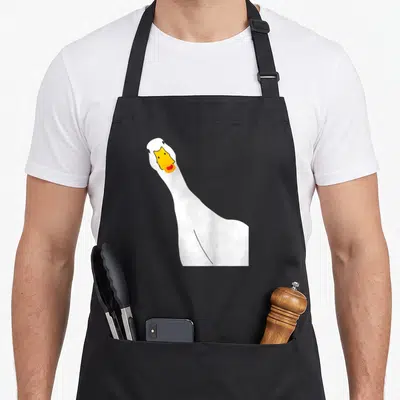Selfie goose apron - Cartoon white goose head and neck with yellow beak and orange mouth detail — shown on a black cotton canvas apron against a white background