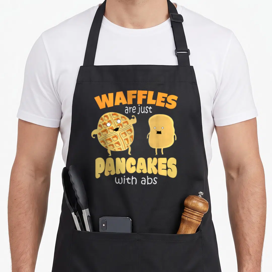Waffle pancake apron - Cartoon waffle flexing arms next to pancake with text WAFFLES are just PANCAKES with abs — shown on a black cotton canvas apron against a white background