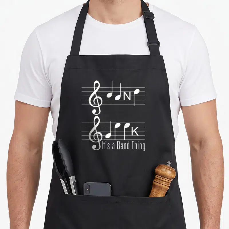 Band geek apron - Two white musical staffs with treble clefs, hidden NK letters, and It's a Band Thing text at bottom — shown on a black cotton canvas apron against a white background