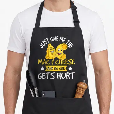 Give mac apron - Cartoon cheese wedge and noodle with Just Give Me The Mac & Cheese text and yellow ribbon — shown on a black cotton canvas apron against a white background