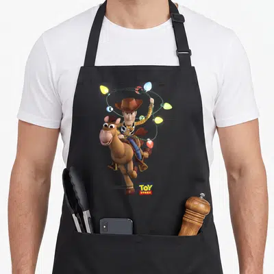 Bullseye camping apron - Woody riding Bullseye galloping pose holding colorful Christmas lights string — shown on a black cotton canvas apron against a white background