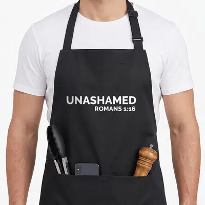 Roman cross apron - White stacked text UNASHAMED ROMANS 1:16 on grey background — shown on a black cotton canvas apron against a white background