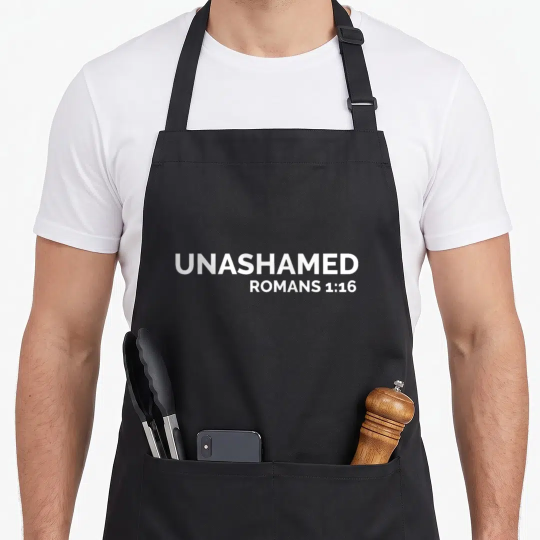 Roman cross apron - White stacked text UNASHAMED ROMANS 1:16 on grey background — shown on a black cotton canvas apron against a white background