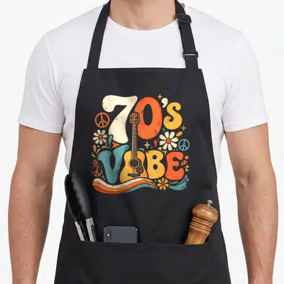 Disco hippie 70s apron - Retro '70's VIBE' text with vertical guitar, peace signs, and daisies in orange, yellow — shown on a black cotton canvas apron against a white background