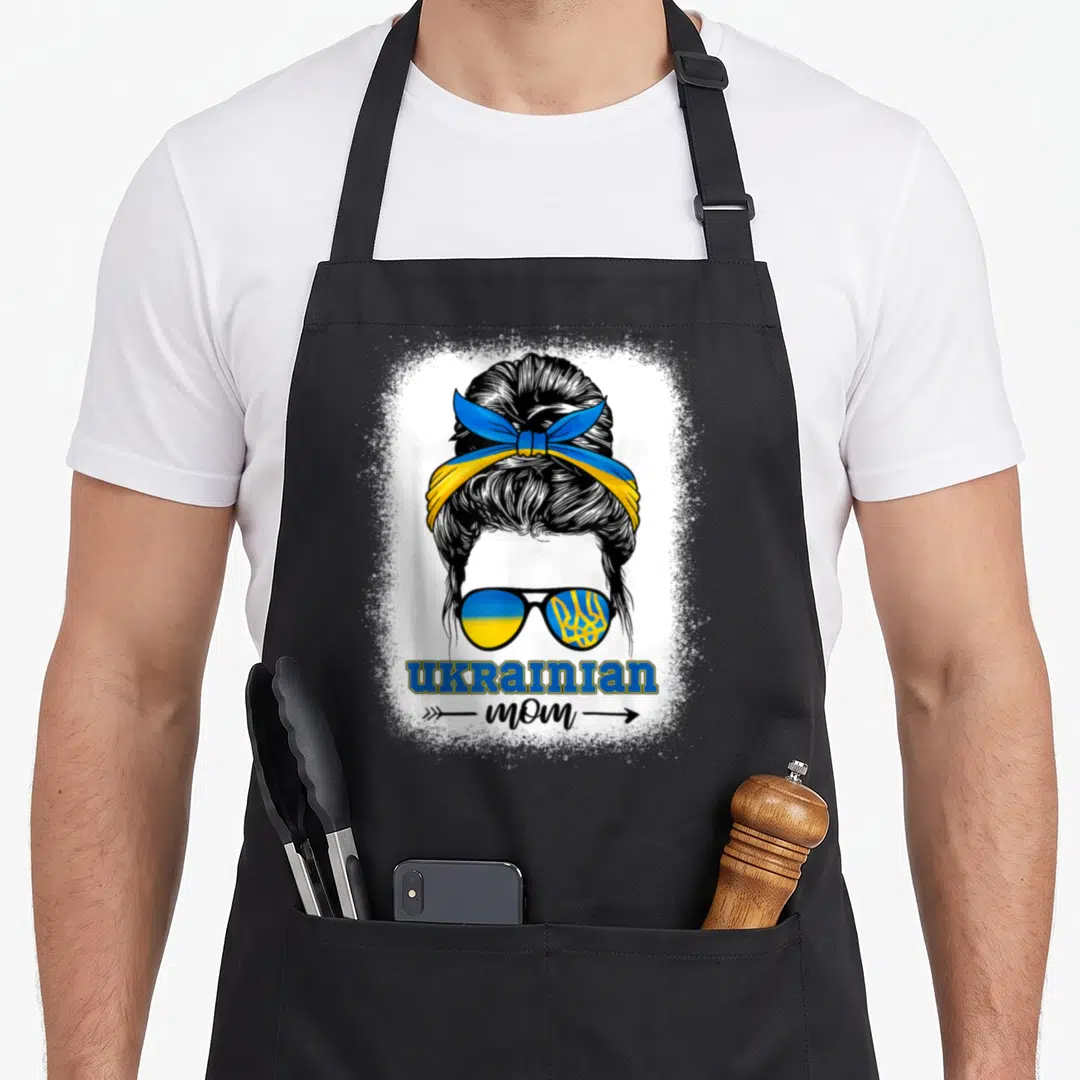 Heritage ukrainian aprons - Distressed woman bun sunglasses blue yellow UKRAINIAN mom text arrows white splatter — shown on a black cotton canvas apron against a white background
