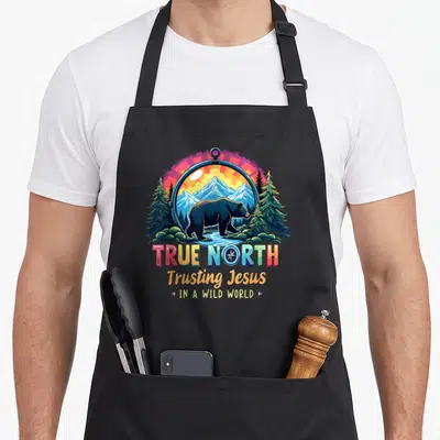 North christian apron - Cartoon bear in compass with mountains and TRUE NORTH Trusting Jesus text — shown on a black cotton canvas apron against a white background