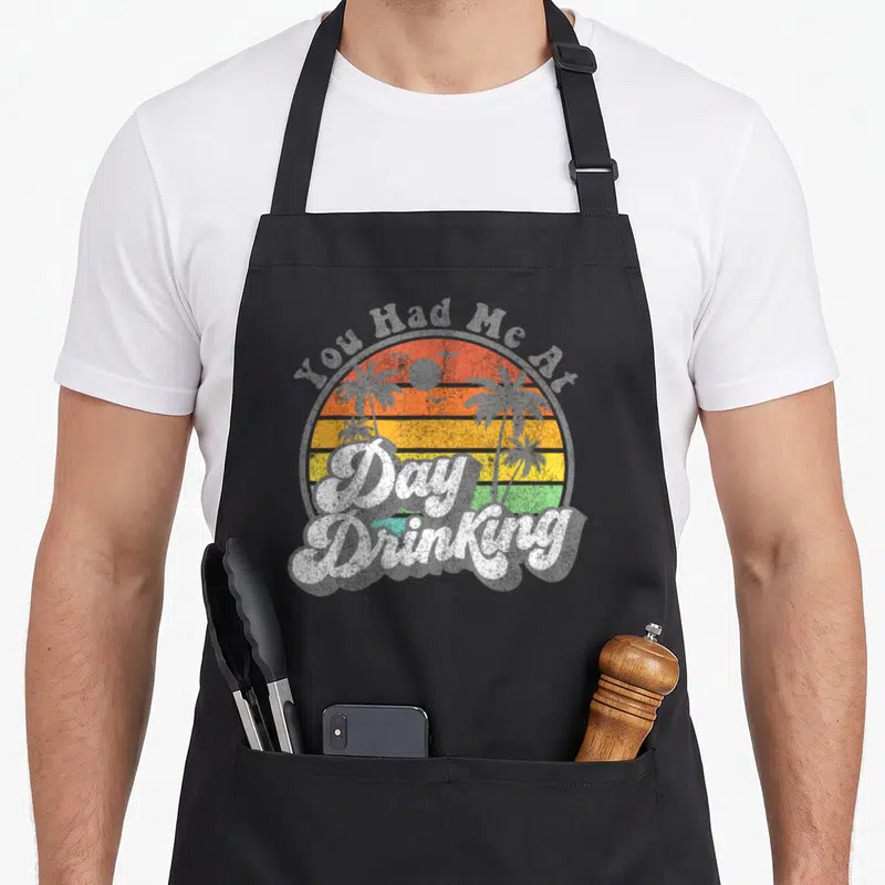 Drinking summer aprons - Retro sunset circle with palm trees and You Had Me Day Drinking text in orange and yellow — shown on a black cotton canvas apron against a white background