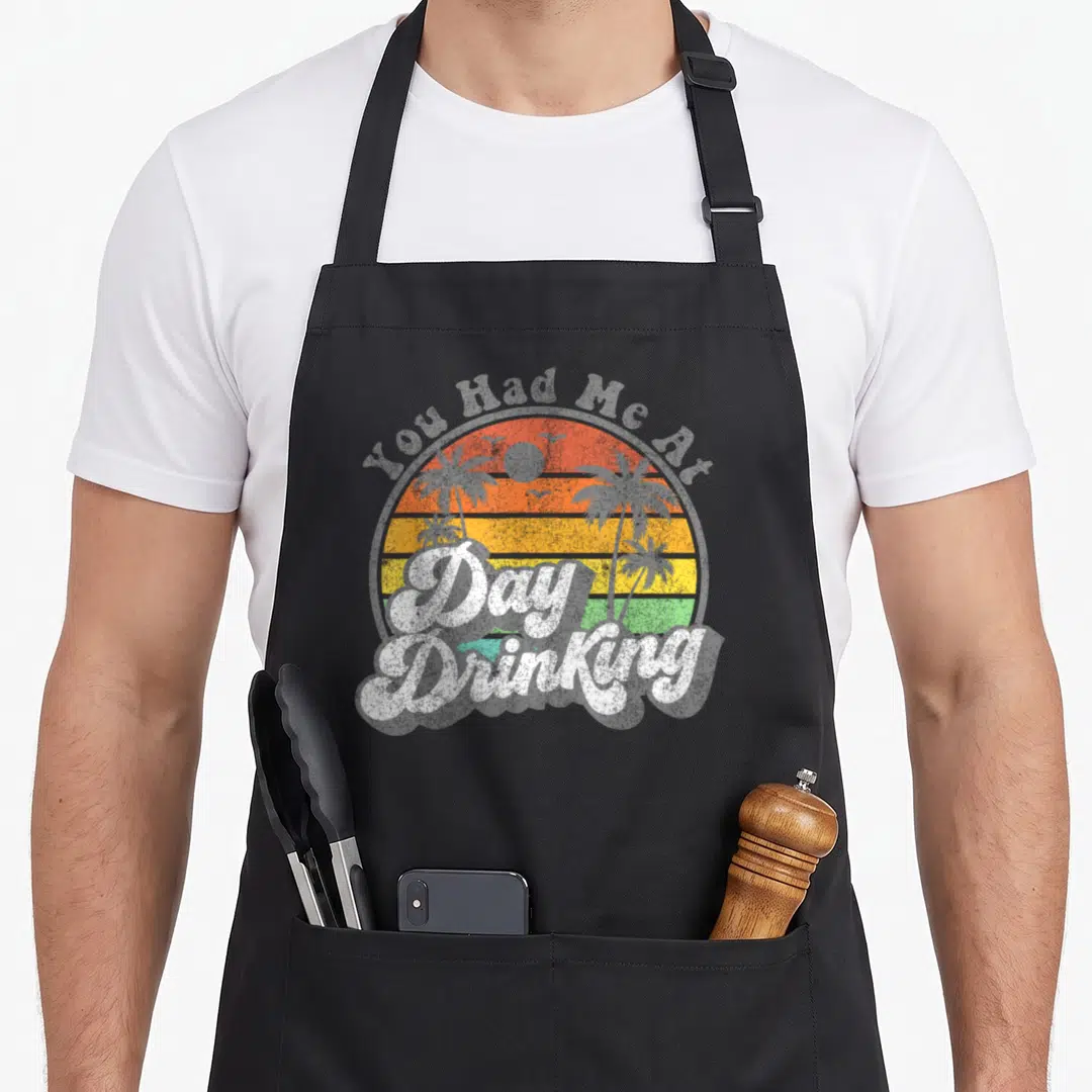 Drinking summer aprons - Retro sunset circle with palm trees and You Had Me Day Drinking text in orange and yellow — shown on a black cotton canvas apron against a white background