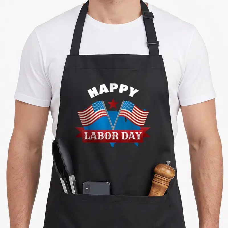 Cute labor apron - HAPPY LABOR DAY text over blue USA map with two crossed red white and blue flags — shown on a black cotton canvas apron against a white background