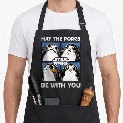 Porg may apron - Four porg heads in 2x2 grid with Star Wars logo and May The Porgs Be With You text — shown on a black cotton canvas apron against a white background