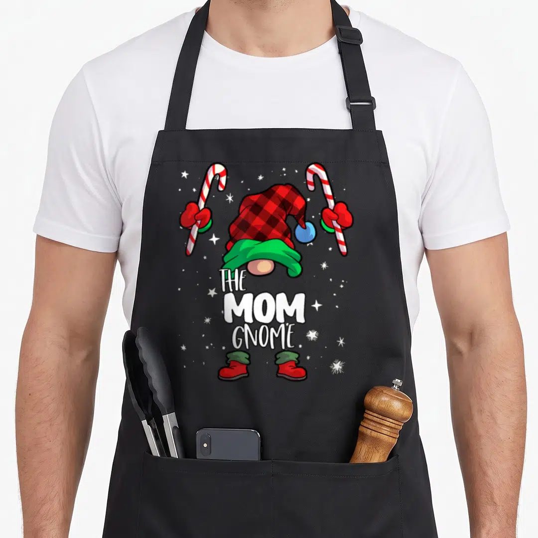 Funny gnome buffalo plaid christmas apron - Cartoon gnome split by THE MOM GNOME text holding candy canes with red — shown on a black cotton canvas apron against a white background