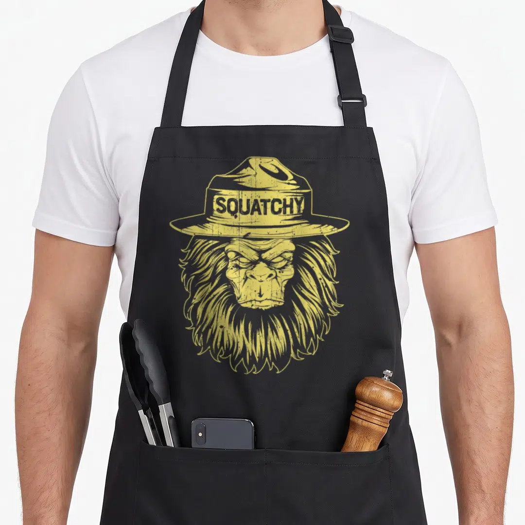 Squatchy bigfoot apron - Yellow distressed bigfoot head wearing a wide-brimmed hat with SQUATCHY text on the band — shown on a black cotton canvas apron against a white background