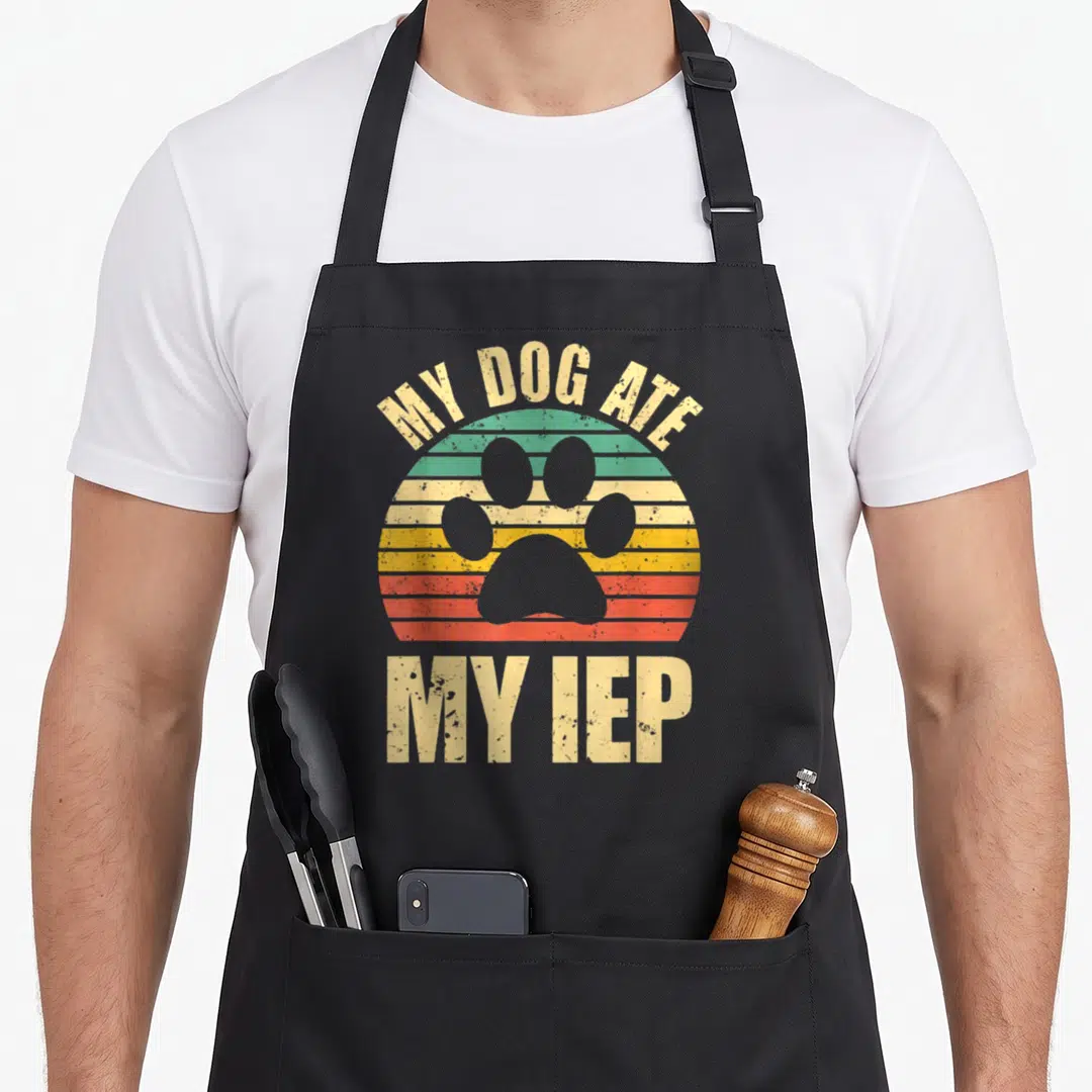 Ate dog apron - Retro sun with paw print and text MY DOG ATE MY IEP — shown on a black cotton canvas apron against a white background