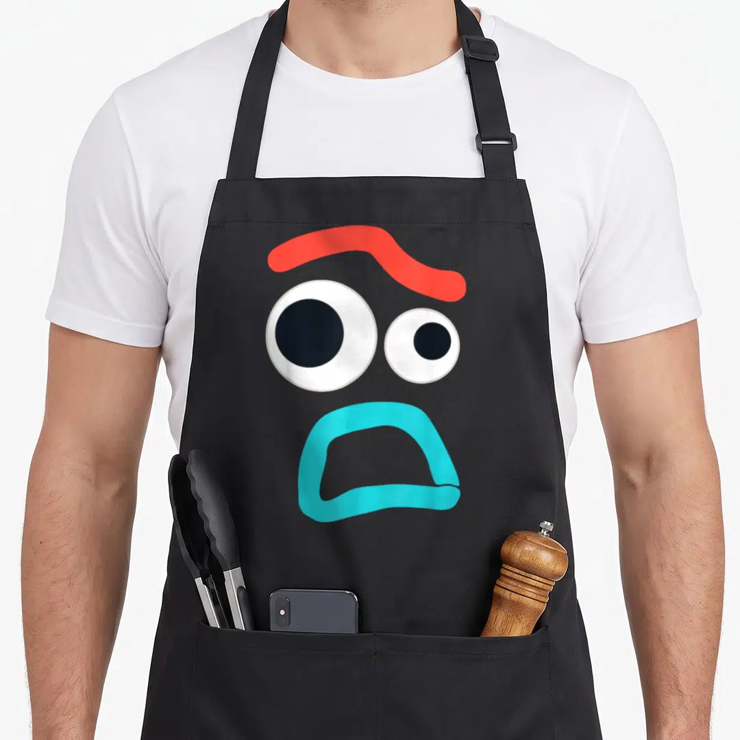 Timid camping apron - Cartoon face with red eyebrow, two white eyes, and light blue mouth on black fabric — shown on a black cotton canvas apron against a white background