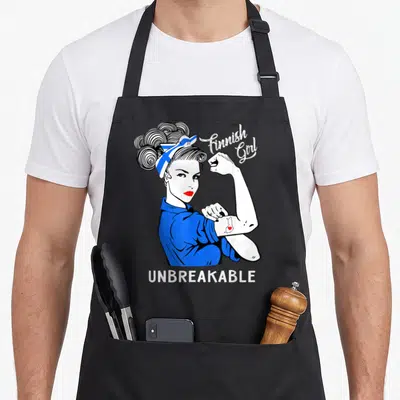 Finnish heritage aprons - Cartoon woman flexing arm wearing Finnish flag headscarf with UNBREAKABLE text below — shown on a black cotton canvas apron against a white background