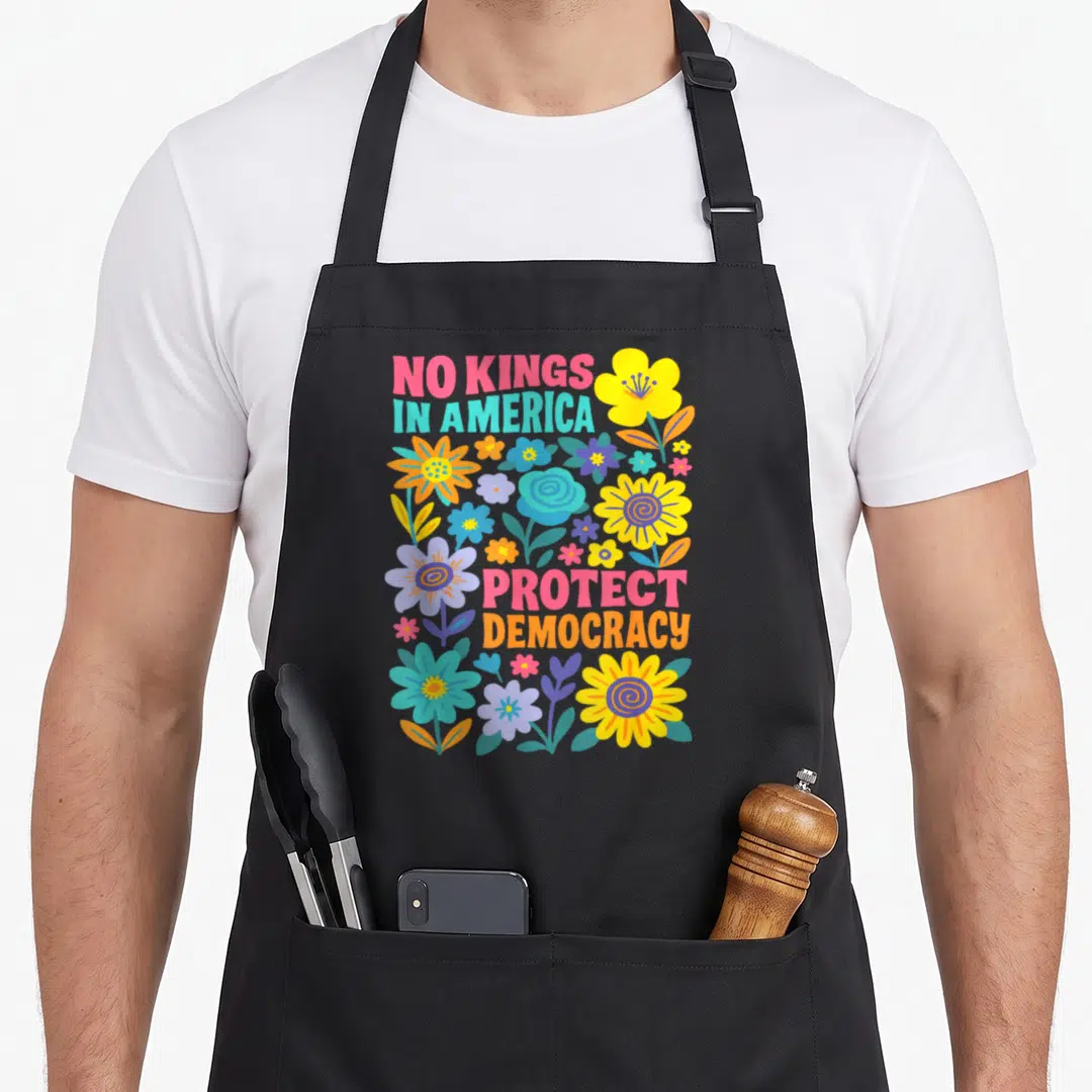 Protest king apron - NO KINGS IN AMERICA PROTECT DEMOCRACY text flanked by pink blue yellow green purple cartoon flowers — shown on a black cotton canvas apron against a white background