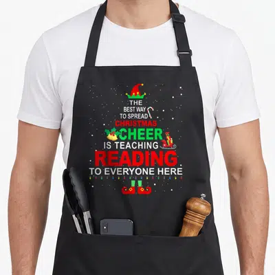 Reading christmas elf apron - Stacked text about teaching reading with Santa hat, elf shoes, candy cane, bells, sleigh — shown on a black cotton canvas apron against a white background